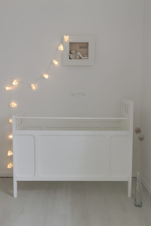 Lit de bébé vintage restauré en blanc en bois. Présenté avec des fleurs séchées et une guirlande lumineuse de coeurs. Univers enfant