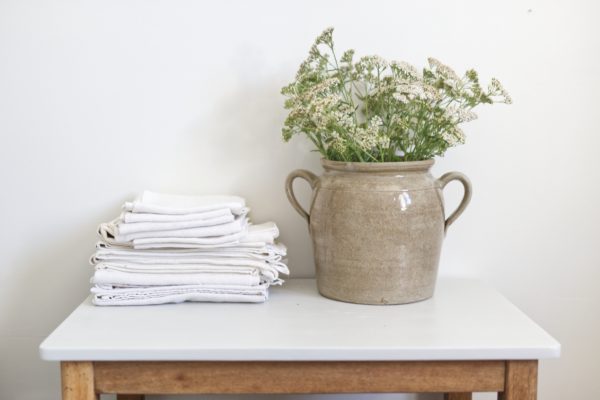 Pot en grès ancien avec des anses servant de vase pour des fleurs sauvages blanches. Tissu ancien blanc sur une table vintage en bois et gris