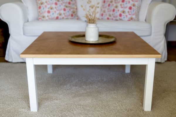 Table basse carrée dont le plateau est en bois et le sous-bassement et les pieds sont en blanc. Canapé blanc, plateau marocain en bronze et pot à moutarde en grès vintage avec des fleurs oreilles de chats séchés