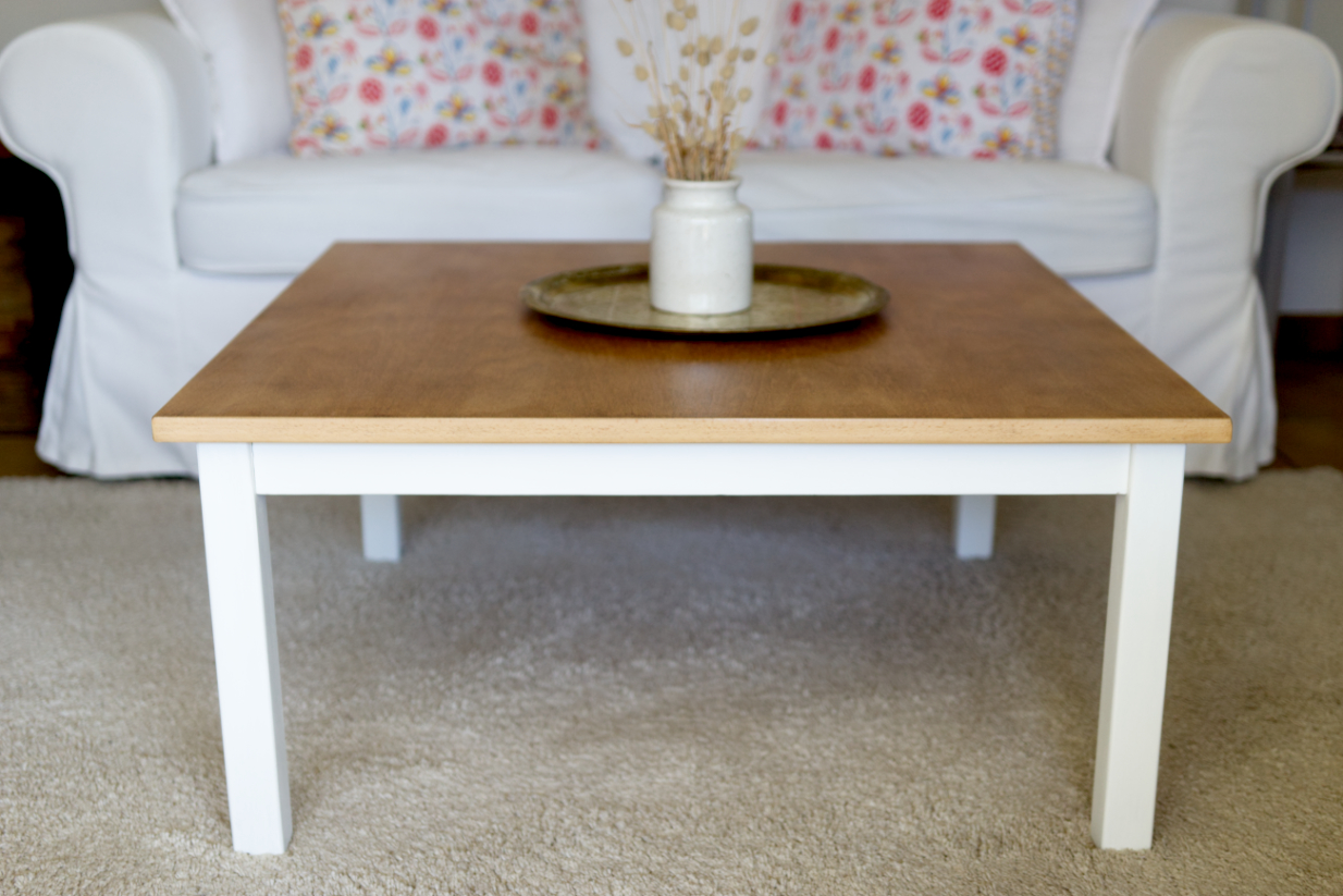 Table basse carrée dont le plateau est en bois et le sous-bassement et les pieds sont en blanc. Canapé blanc, plateau marocain en bronze et pot à moutarde en grès vintage avec des fleurs oreilles de chats séchés