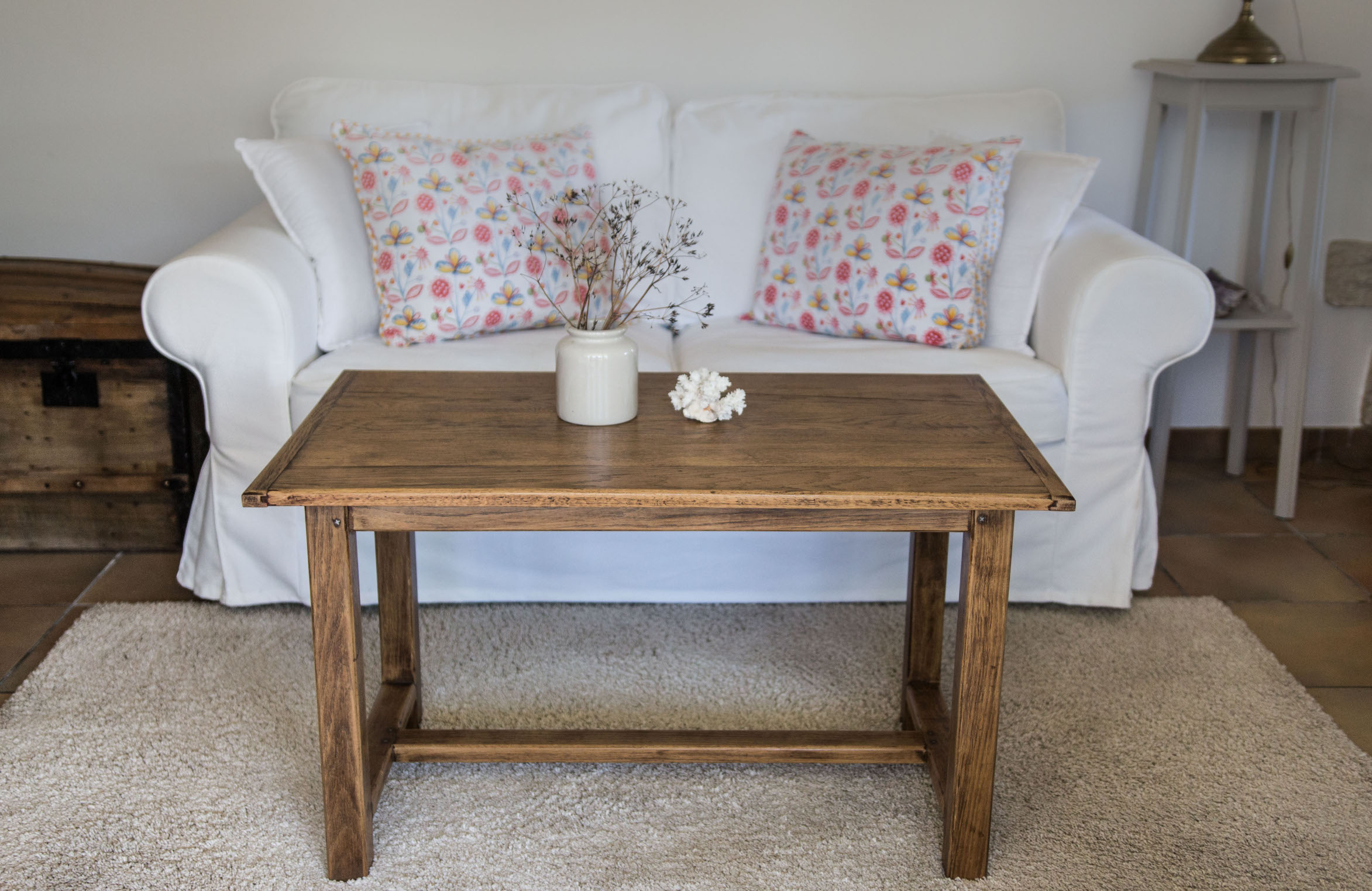 Table en bois brut rénovée vintage poncée et vernie. Canapé blanc, malle ancienne, corail et fleurs séchées dans un pot à moutarde en grès ancien